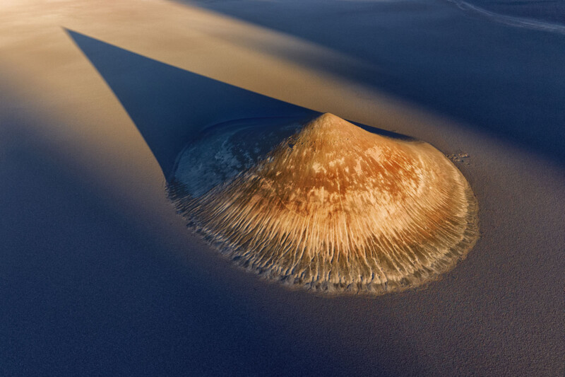 Aerial view of a conical sand dune casting a long, sharp shadow across a flat, barren desert landscape, with textured ridges radiating from the base of the dune.