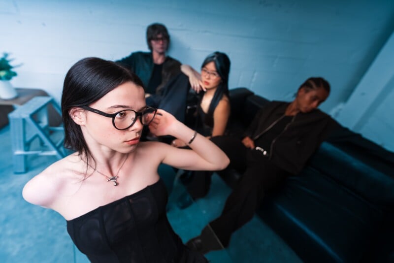 A young woman with glasses poses in the foreground, adjusting her hair, while three people dressed in black sit on a dark sofa in the background of a modern, blue-toned room.