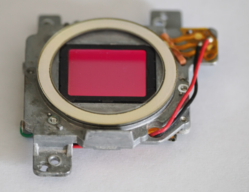 A close-up of a digital camera sensor assembly featuring a metallic frame, visible wiring, and a prominent red rectangular sensor in the center.
