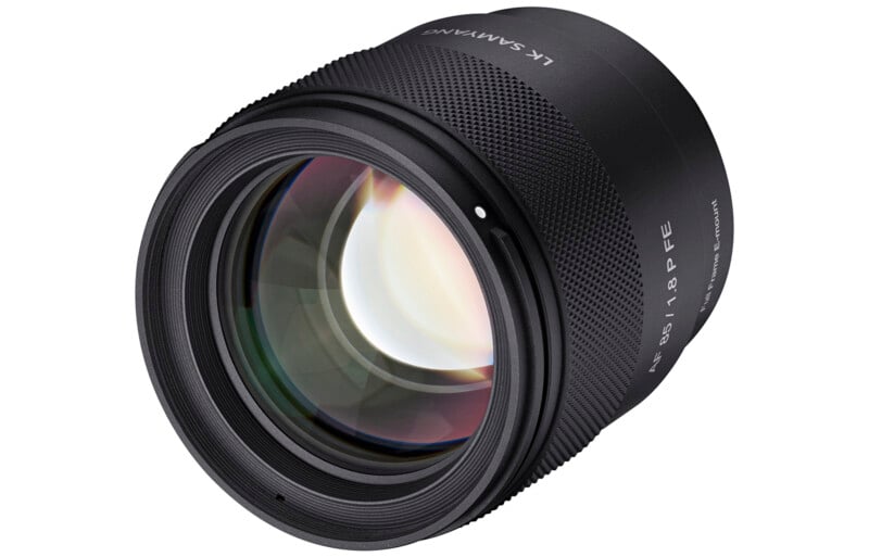 A close-up of a black Samyang camera lens with visible glass elements and text markings indicating it is an AF 85mm F1.4 FE model. The lens is angled against a white background.