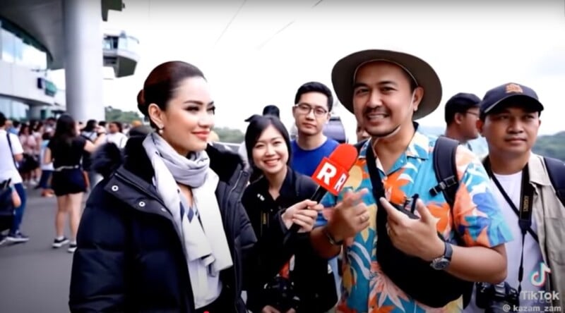 A woman holding a red microphone interviews a smiling man in a floral shirt and hat, joined by three other people standing outdoors in a crowded area.