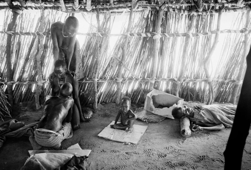 Three adults and a baby are inside a hut made of vertical wooden sticks. One adult interacts with the baby on a mat, another sits nearby, and a third lies on the ground, partially covered by a blanket. Sunlight filters through the walls.