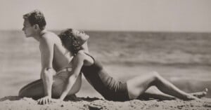 A man and woman in vintage swimsuits sit back to back on a sandy beach, facing the ocean. The woman leans back with her face tilted upward, eyes closed, enjoying the sun. The photo is in black and white.