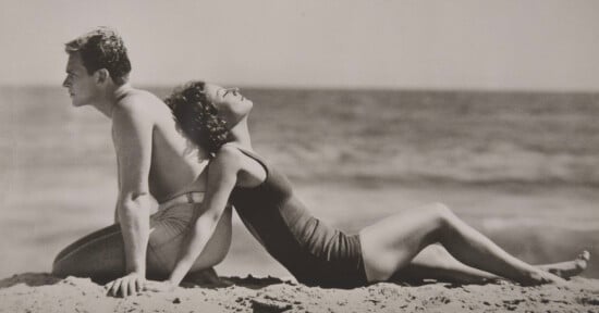 A man and woman in vintage swimsuits sit back to back on a sandy beach, facing the ocean. The woman leans back with her face tilted upward, eyes closed, enjoying the sun. The photo is in black and white.
