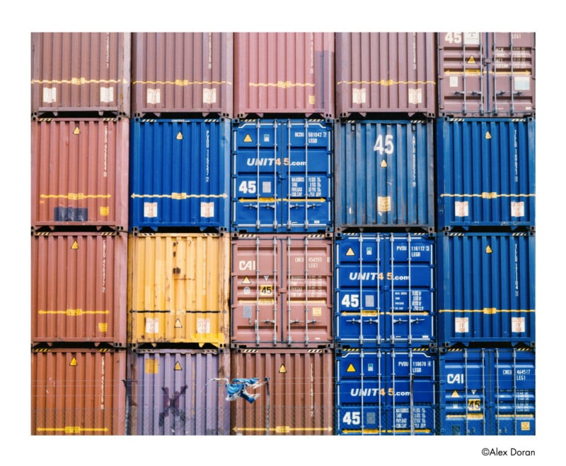 A wall of stacked shipping containers in blue, yellow, and brown hues. A person on a bicycle rides in front of the containers, adding a sense of scale to the scene.