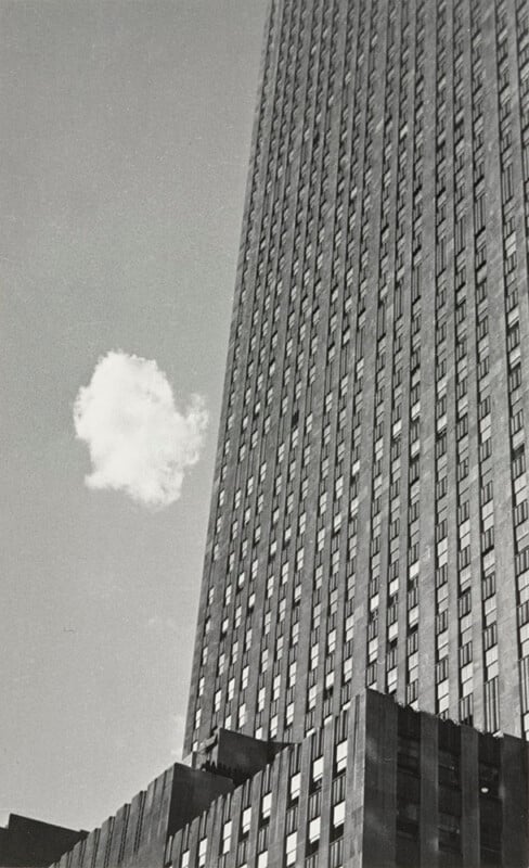 A single small cloud floats near the top of a tall, modern skyscraper with many windows, photographed from a low angle against a clear sky.