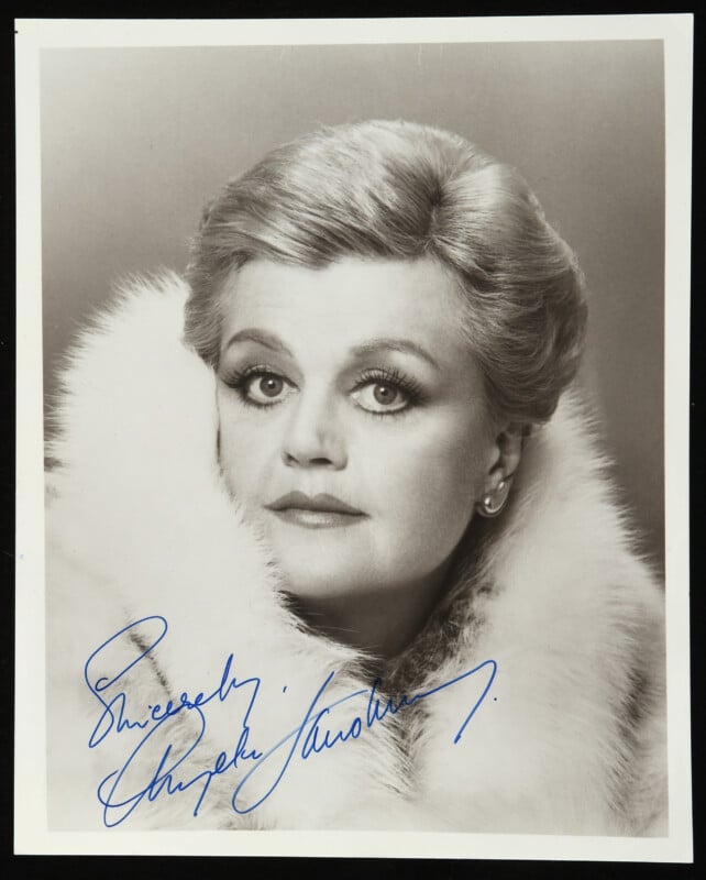 A black-and-white portrait of a woman with styled light hair, wearing a fur wrap, looking directly at the camera. The photo is signed "Sincerely, Angela Lansbury" in blue ink.