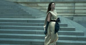 A woman in a cream satin dress with a large black bow and long black gloves stands near wide stone steps, gazing pensively to the side. The background features a grand, gray stone building.