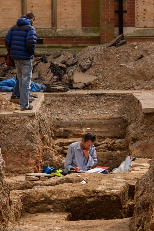 A person sits and writes notes in an archaeological excavation trench while another person in a blue jacket stands nearby, looking at the site. Dirt, rocks, and building rubble surround them.