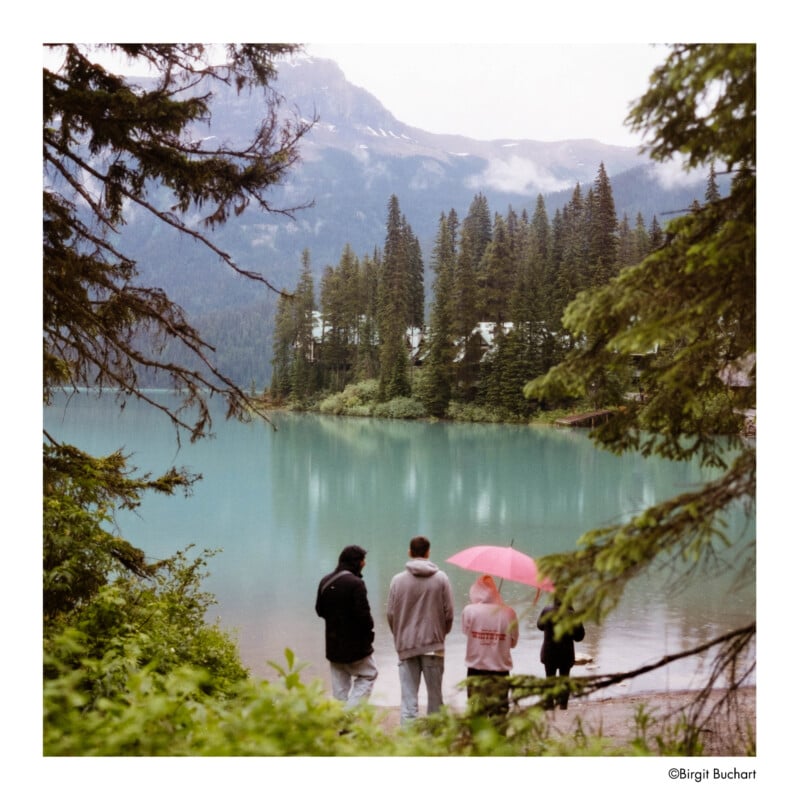 Four people stand on the shore of a blue-green lake surrounded by pine trees and mountains; one person holds a pink umbrella. The sky is cloudy, and the scene is framed by tree branches.