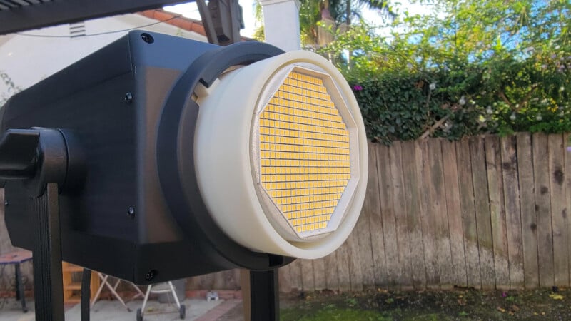 A close-up of a studio LED light with a hexagonal grid diffuser, set up outdoors near a wooden fence and greenery in the background.