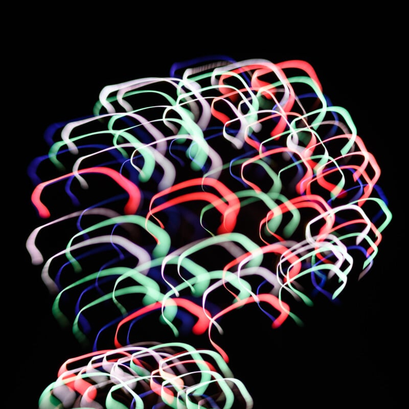 Colorful light trails create overlapping, abstract loops in red, green, blue, and white on a black background, forming a vibrant, dynamic pattern resembling fireworks or neon shapes.