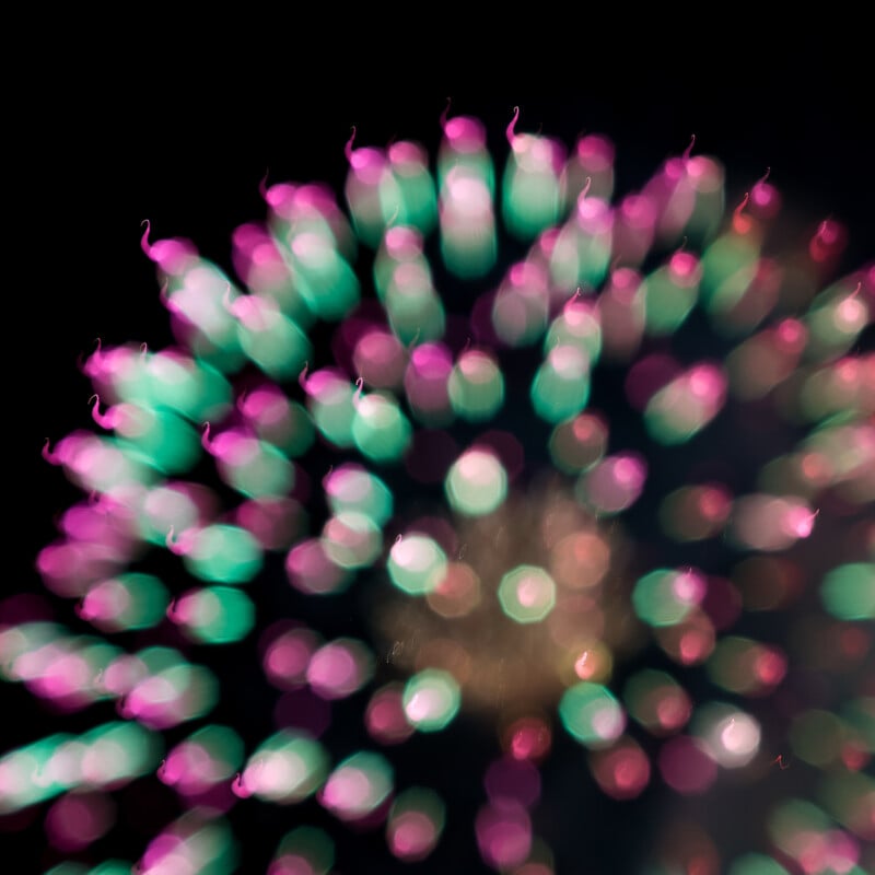 A colorful, abstract view of fireworks against a black sky, with green and pink light bursts forming a circular pattern. The image is intentionally out of focus, creating a soft, dreamy effect.