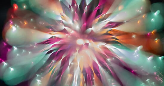 A vibrant, abstract burst of colors including teal, pink, orange, and white, radiating outward from a central bright point, resembling an explosion or a firework viewed through a soft, dreamy lens.