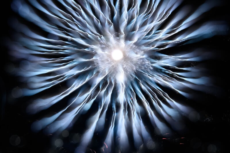 A burst of white and blue light streaks radiates outward from a bright central point against a dark background, resembling an abstract firework or energy explosion.