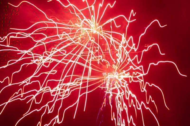 Bright orange and yellow fireworks streak across a red-lit sky at night, creating swirling, wavy patterns of light and energy in the darkness.