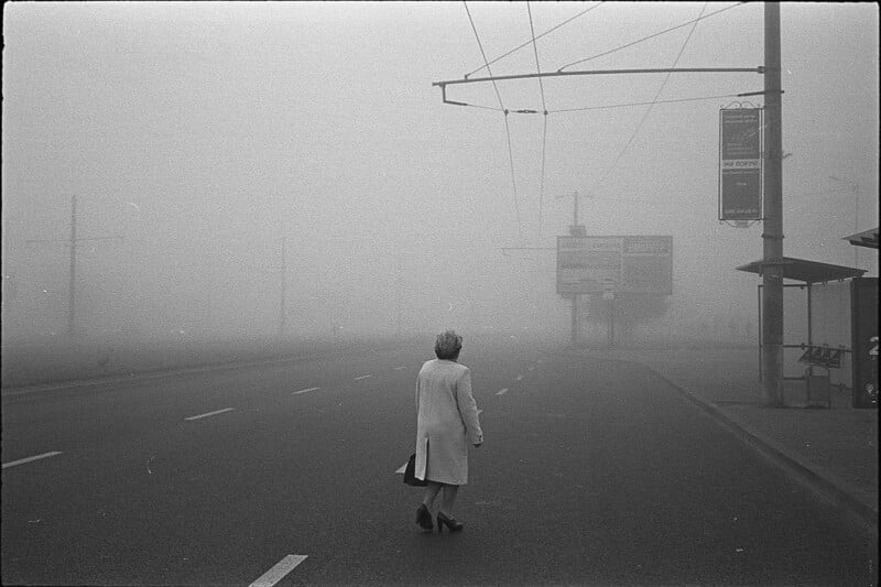 A person wearing a light-colored coat walks alone across an empty street enveloped in thick fog, making the surroundings, street signs, and buildings barely visible.