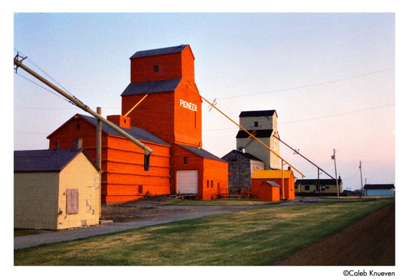 A row of brightly colored grain elevators, including a prominent orange one labeled "Pioneer," stands beside a rural road at sunset. Grassy areas and utility poles are visible alongside the buildings.