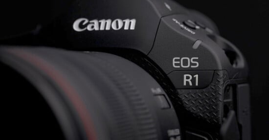 Close-up of a black Canon EOS R1 camera, showing the Canon brand logo and the model name “EOS R1” near the textured grip, with part of the lens visible on the left.