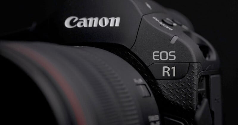 Close-up of a black Canon EOS R1 camera, showing the Canon brand logo and the model name “EOS R1” near the textured grip, with part of the lens visible on the left.