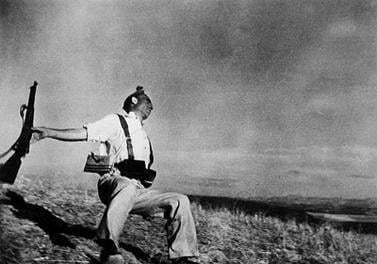 A man in military attire is captured mid-fall on a grassy slope, his head tilted back and arms outstretched, holding a rifle. The scene conveys a sense of sudden movement and dramatic tension under a wide, open sky.