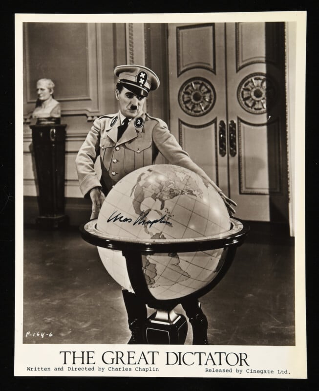 Black-and-white photo of Charlie Chaplin in a military uniform as his character in "The Great Dictator," standing with hands on a large globe, with ornate doors and a bust visible in the background.