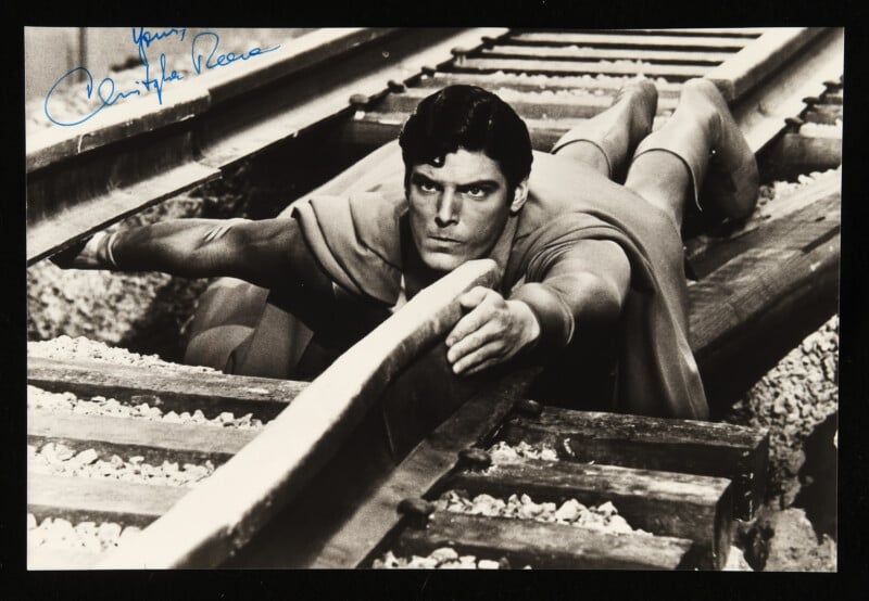 A man dressed as Superman lies between railroad tracks, holding the rails apart with his hands and shoulders to stop a potential train accident. The photo is black and white and has an autograph in the top left corner.
