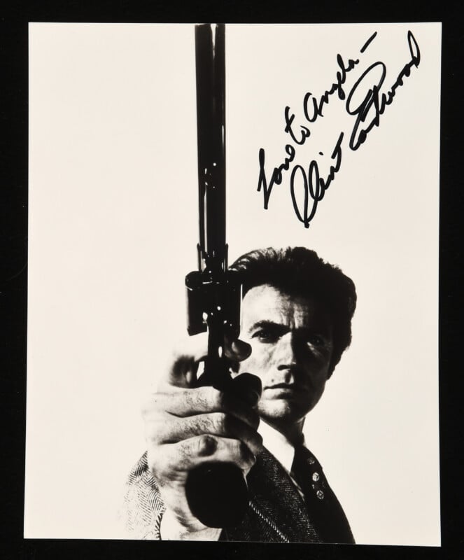 A man in a suit points a large revolver directly at the camera, with his face partially shadowed. The photo is black and white and features a handwritten autograph in the top right corner.