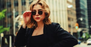 A woman with shoulder-length blonde hair wearing sunglasses and a black outfit stands confidently outdoors in an urban setting, holding her sunglasses with one hand.