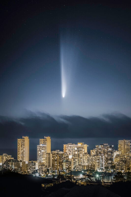 A bright comet with a long glowing tail streaks across the night sky above a city skyline with illuminated high-rise buildings. Thin clouds hover above the horizon, and stars are visible in the sky.