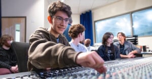 A young person adjusts controls on a large audio mixing console in a recording studio, with several others sitting and talking in the background.