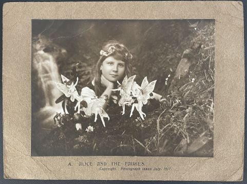 A black-and-white photo of a young girl with a flower crown, posed outdoors beside a group of illustrated fairies among flowers. Text at the bottom reads, "Alice and the Fairies, Copyright Photograph Taken July 1917.