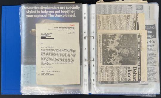 A blue binder holds plastic sleeves containing a typed letter and several newspaper clippings, some with headlines and photos, organized for archival or research purposes.
