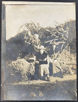 Four women in long dresses and hats sit outdoors on a grassy, bushy hillside. Two are seated higher up and two below, all facing the camera in bright sunlight. The photo appears old and sepia-toned.