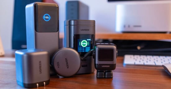 Several UGREEN electronic devices, including chargers and adapters, are displayed on a wooden desk in front of a computer setup, with digital screens showing charging icons.