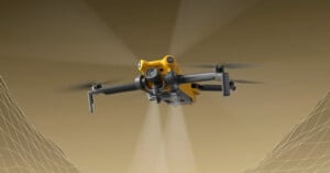 A yellow quadcopter drone with a mounted camera is flying forward against a brown background, with faint digital grid lines representing terrain or mapping data underneath its wings.