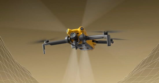 A yellow quadcopter drone with a mounted camera is flying forward against a brown background, with faint digital grid lines representing terrain or mapping data underneath its wings.