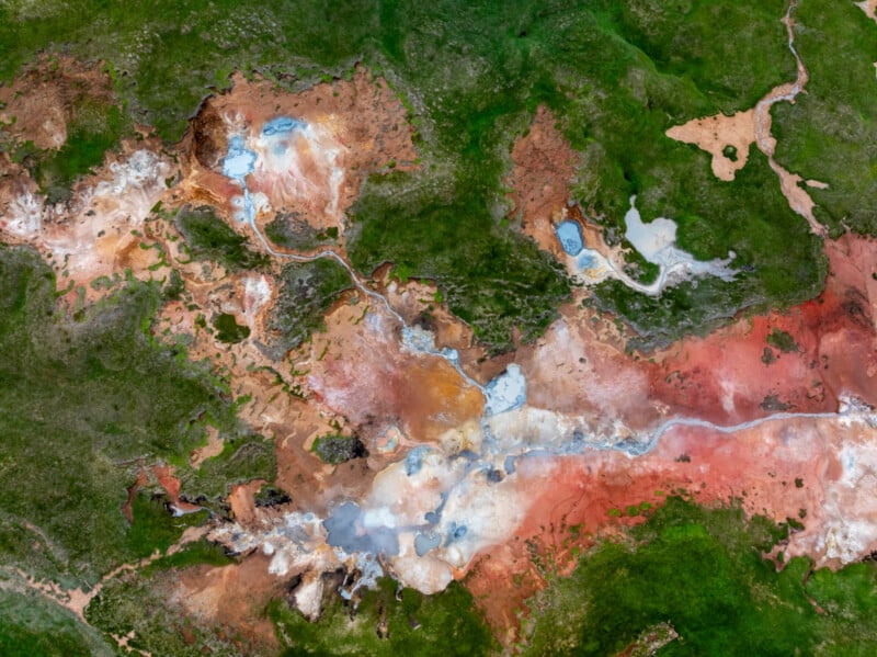 Aerial view of a geothermal landscape with vivid green grass, rust-red soil, white mineral deposits, and small blue pools connected by winding streams. The terrain displays striking natural colors and abstract patterns.