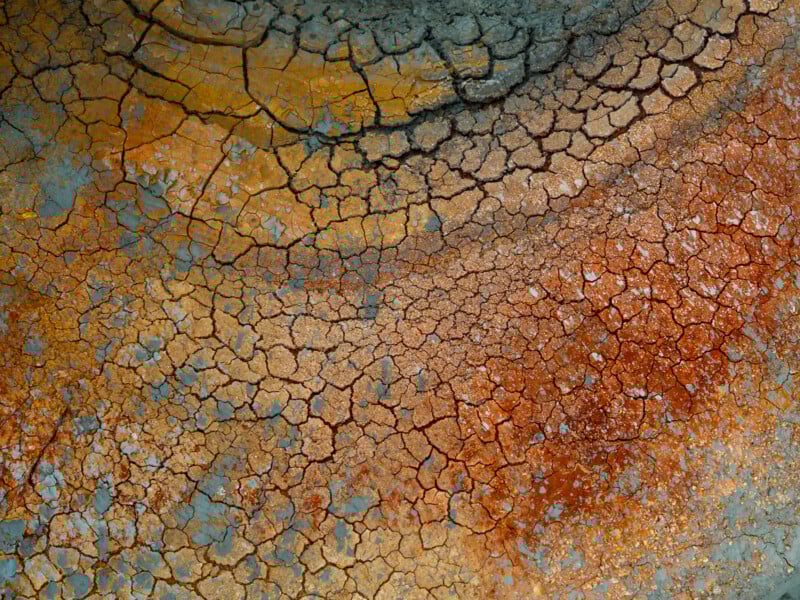 Cracked, dry earth in shades of brown, orange, yellow, and gray, forming an abstract, textured pattern that resembles a parched landscape or dried paint.
