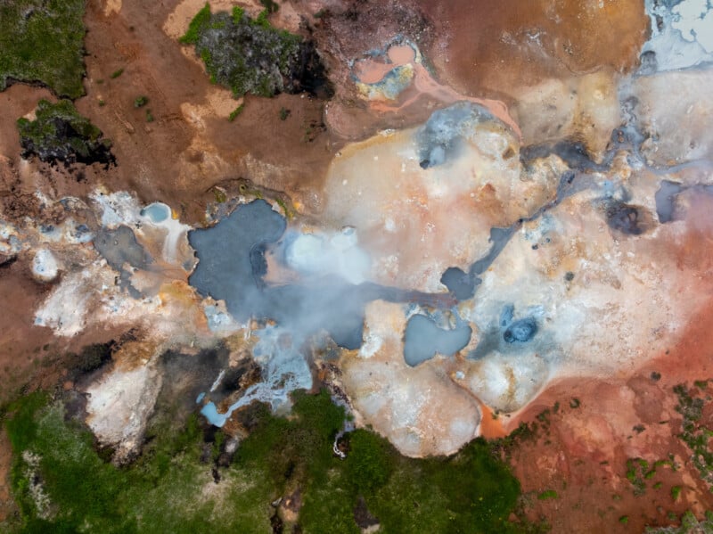 Aerial view of a colorful geothermal hot spring area with steaming pools, mineral deposits in white, orange, and blue hues, and patches of green vegetation at the edges.