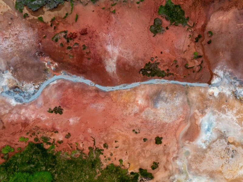 Aerial view of a colorful, textured landscape featuring red, orange, and brown earth with patches of green vegetation and a narrow, winding gray stream running through the center.