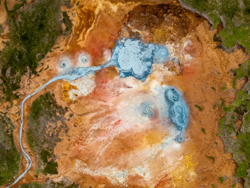 Aerial view of a geothermal hot spring with vivid orange, yellow, and green mineral deposits, blue steaming pools, and surrounding patches of green vegetation.