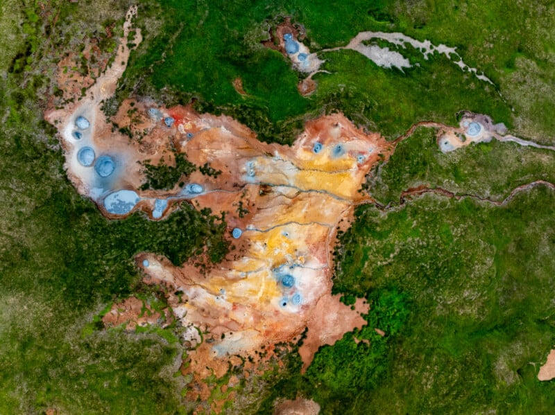 Aerial view of a colorful geothermal area with orange, yellow, and brown mineral deposits, scattered blue hot springs, and green grass surrounding the unique landscape formation.