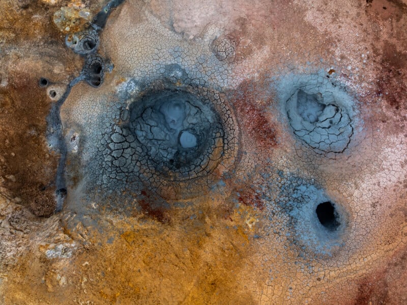 Aerial view of a geothermal area with cracked blue-gray mud pools, surrounded by reddish-brown and yellow earth, showing textured patterns and mineral deposits.