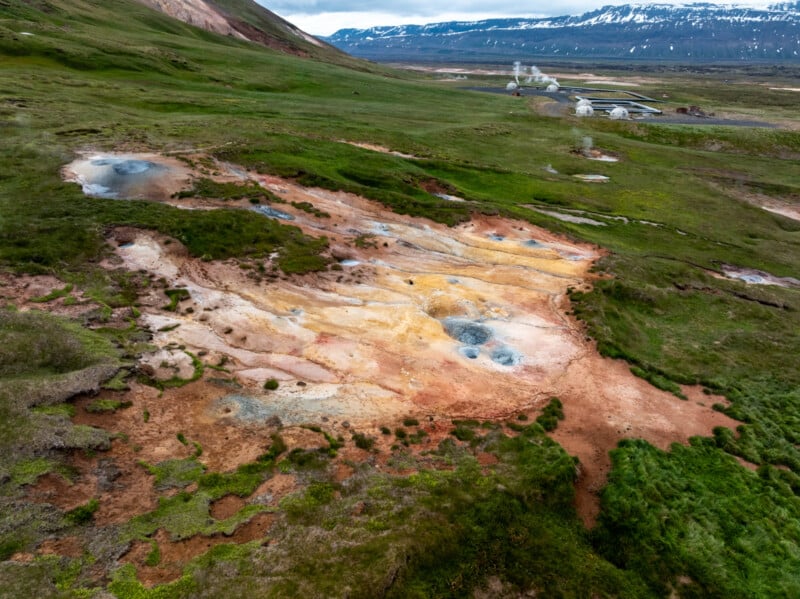 Aerial view of a geothermal area with steaming vents, multicolored earth, patches of green grass, and distant mountains partially covered with snow.