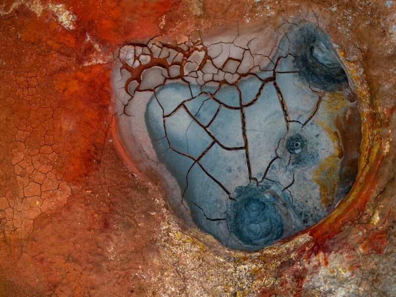 Aerial view of a cracked, dry mud pool surrounded by vibrant red, orange, and beige earth, creating an abstract, textured landscape with natural patterns and color contrasts.