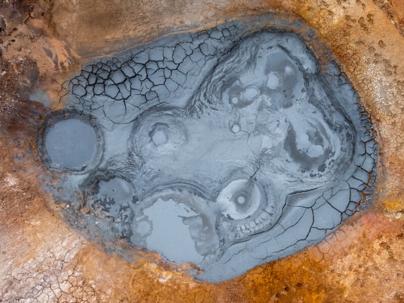 Aerial view of a bubbling mud pool with cracked, dry edges and patches of smooth, grey mud in a geothermal area surrounded by orange and brown earth.