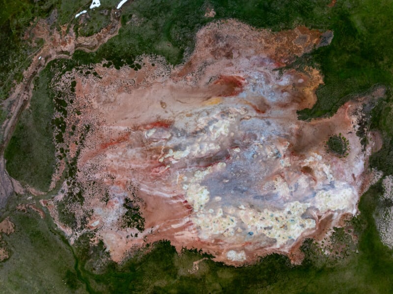 Aerial view of a large, multicolored geothermal area with patches of green vegetation on the edges and varying shades of brown, red, white, yellow, and gray across the surface, creating a textured abstract pattern.