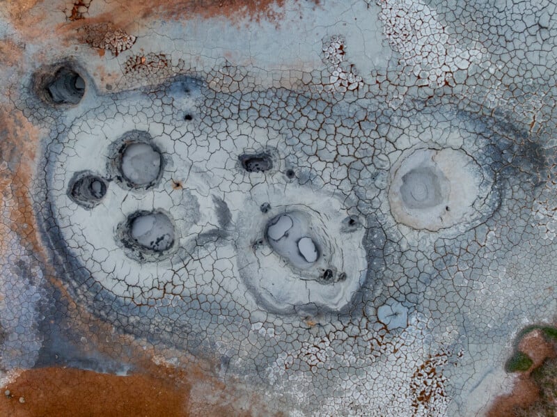 Aerial view of cracked, dry earth with several circular mud pools and patches of white and reddish soil, resembling a geothermal or volcanic landscape.