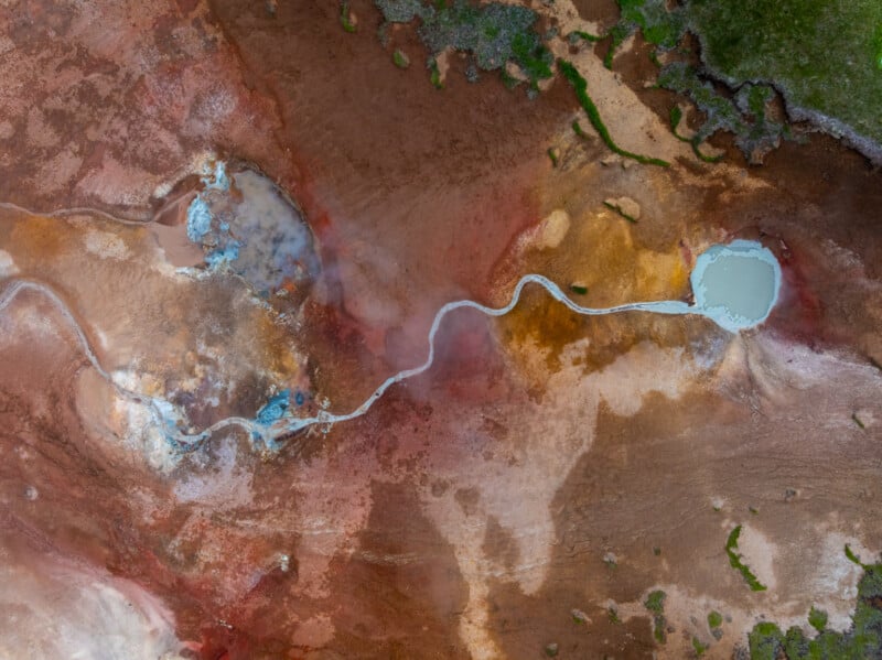 Aerial view of a geothermal area with colorful earth tones, showing a winding white path connecting bubbling pools of water, surrounded by green patches of vegetation.